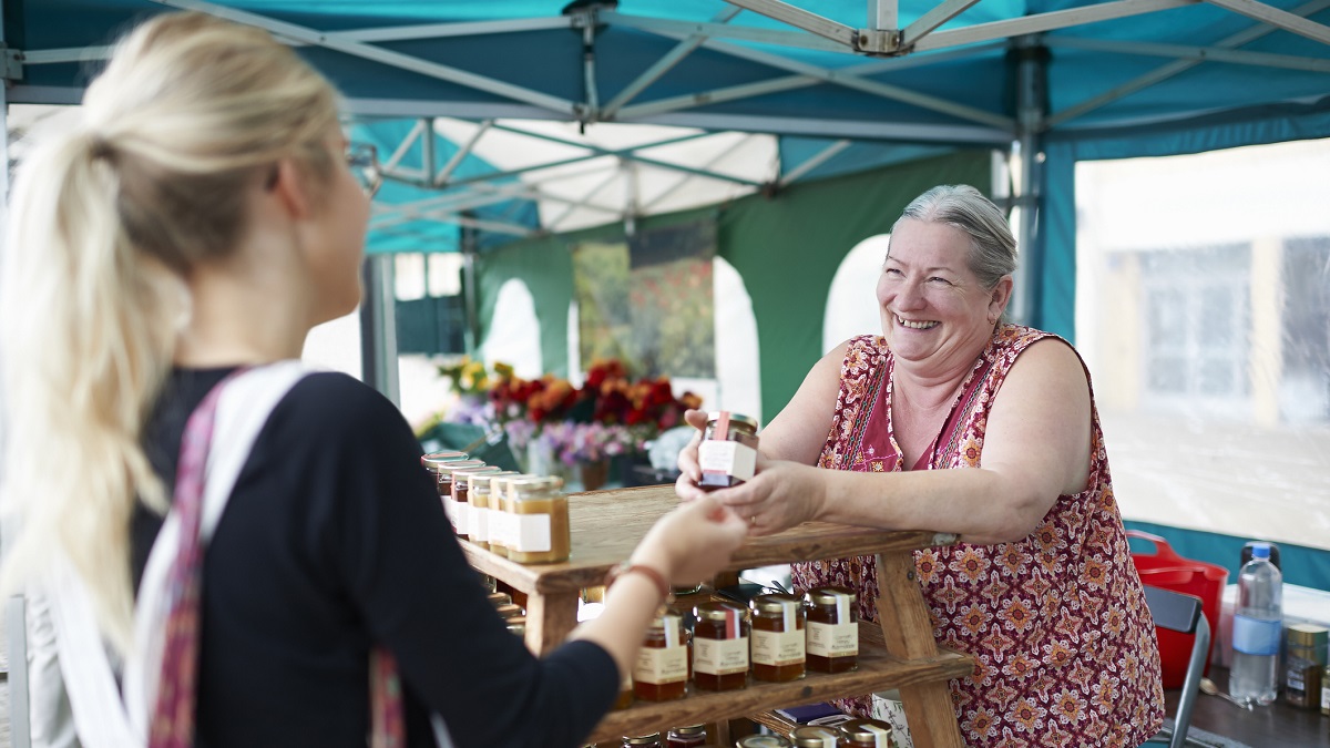 A senior woman working on a local market stall selling homemade jam serves a customer.