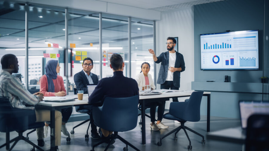 Diverse Modern Office: Businessman Leads Business Meeting With Managers, Talks, Uses Presentation Tv With Statistics, Infographics. Digital Entrepreneurs Work On E Commerce Project.